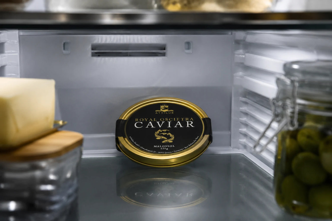 A sealed tin of Attilus Royal Oscietra Caviar resting on a refrigerator shelf alongside a wedge of cheese and a jar of green olives, a refined home-storage scene.