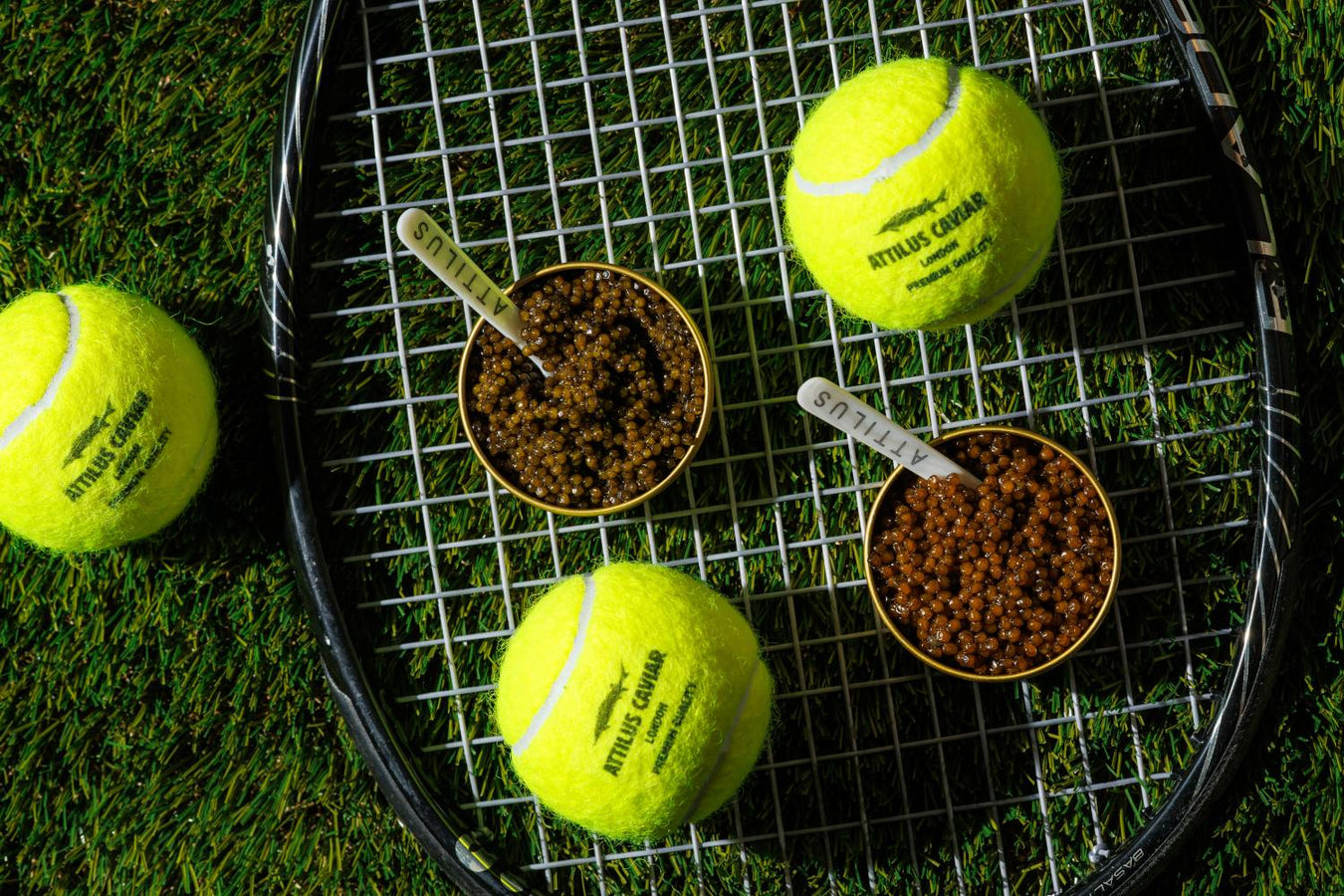 Two open tins of Attilus caviar with branded spoons rest on a tennis racquet alongside Attilus-branded tennis balls on grass, an editorial Wimbledon-style scene.